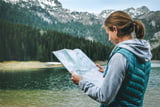 Eine junge Frau steht am Rande eines grünen Sees in den Bergen, trägt eine grüne Weste und hat eine Wanderkarte in der Hand.
