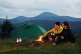 Ein junges Pärchen sitzt vor seinem Zelt in den Bergen am Lagerfeuer und hält Marshmellows in die Flammen.