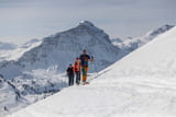 Mehrere Personen sind auf Tourenski in einer verschneiten Berglandschaft bei sonnigem Wetter im Aufstieg unterwegs.