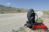 Am Rand einer staubigen landstraße in Kirgistan steht ein Trekkingrucksack, Wanderschuhe und Wanderstöcke für eine Trekking-Tour bereit.