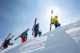 Vier junge Personen sind auf einem Gipfelgrat unterwegs. An ihre Lawinenrucksäcke haben sie ihre Tourenskier geschnallt, in der Hand haben sie jeweils rechts einen Skitourenstock und rechts einen Eispickel.